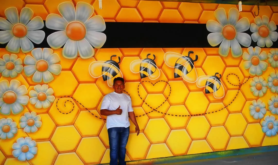 Mural amarelo com colmeia, flores e abelhas, com o artista.