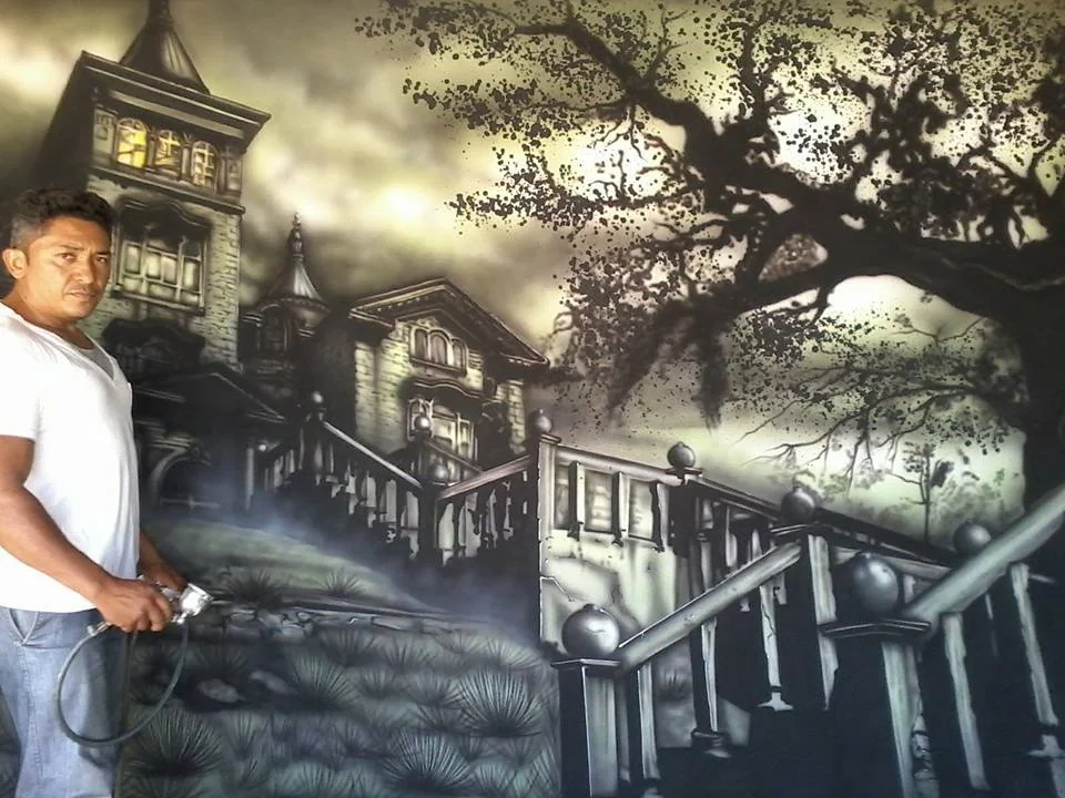 Mural monocromático com casas e árvores; o artista segura a pistola de pintura.